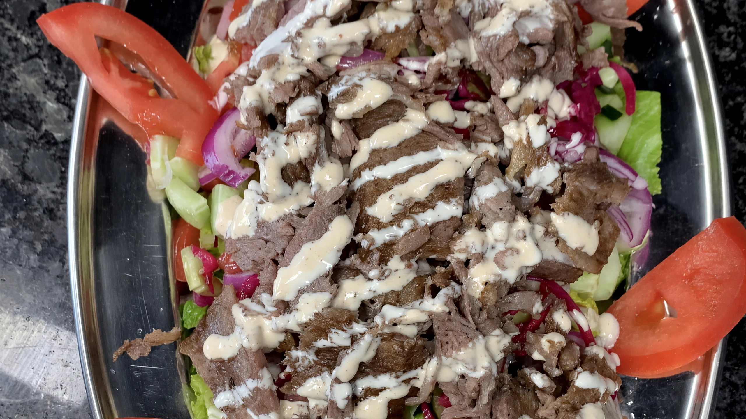 Sliced beef gyro over salad with garlic sauce and tomato on an oval steel platter